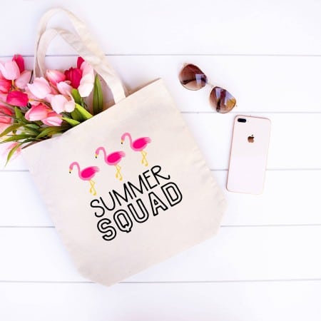 Canvas tote bag on a white shiplap background holding flowers, with sunglasses and a phone nearby. The totebag has an image saying Summer Squad with a group of flamingos.