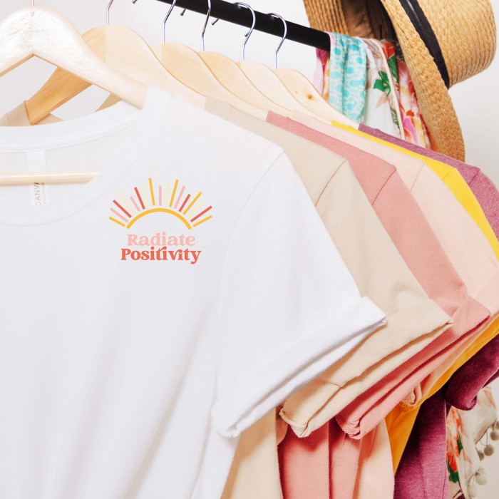 many shirts of various colors hanging on a clothing rack. The first shirt is white and has a svg file design showing arrays of sunshine and radiate positivity written in vintage style text