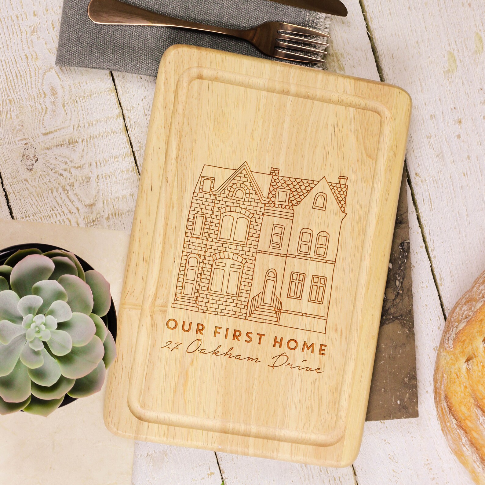a custom cutting board that has a hand drawn image of a home with the text Our First Home and the address listed underneath it. This is a great glowforge project to make and sell.