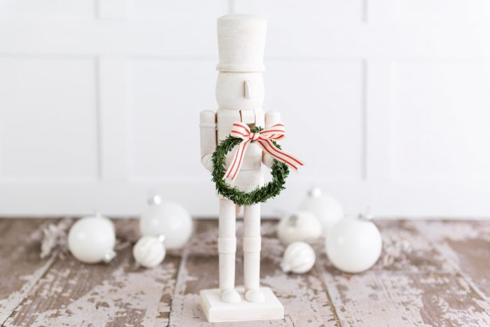 Farmhouse modern style nutcracker painted all white and holding a green wreath with a ribbon bow.