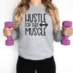 A woman is holding purple hand weights in a grey crewneck sweatshirt that says Hustle For That Muscle with an image of a weight.