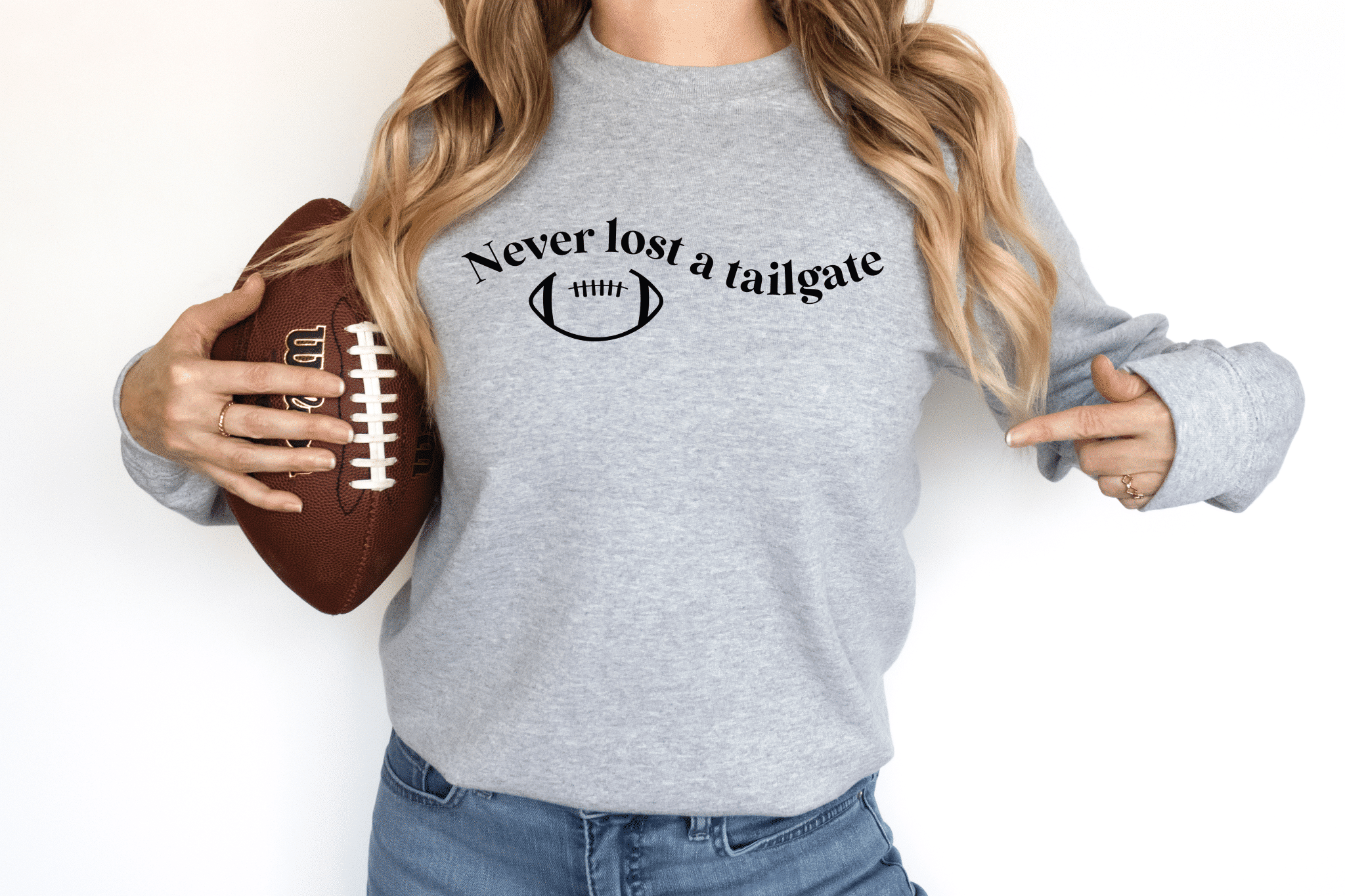 Woman holding a football wearing a grey crewneck sweatshirt that says never lost a tailgate