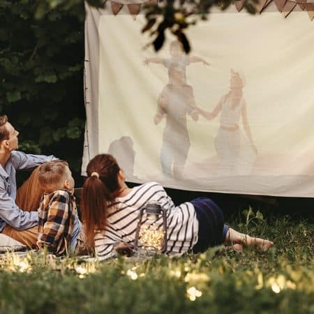 Parents and son sitting on blanket with pillows and watching movie projected on cloth in summer evening in yard