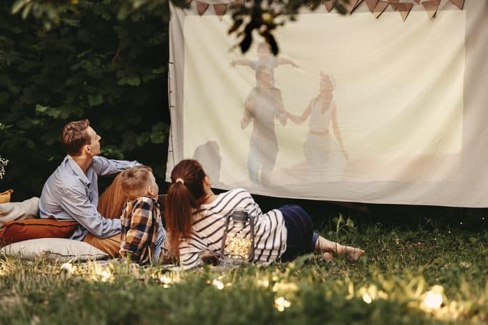 Parents and son sitting on blanket with pillows and watching movie projected on cloth as a diy outdoor move theater.